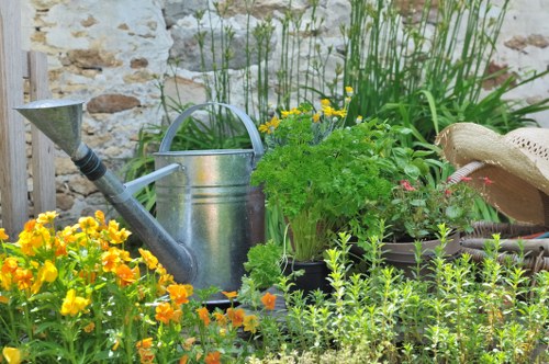 The image depicts a small, lush garden area featuring a variety of plants and garden elements. In the foreground, there are vibrant yellow and orange flowering plants, possibly primroses, with dense green foliage. Behind these, a collection of potted herbs and decorative plants are arranged, including a leafy green herb plant. To the left, a traditional metal watering can with a rounded handle and long spout is placed among the plants, indicating outdoor maintenance activity. Adjacent to the watering can, a wooden garden feature or border is partially visible, with other garden tools or accessories nearby. The environment appears to be a cultivated outdoor space, possibly part of a private garden, with a stone or brick wall serving as a backdrop, suggesting a well-maintained and thoughtfully arranged garden setup. The lighting suggests natural daylight with no harsh shadows, indicative of fair weather conditions typical of outdoor gardening in London or nearby areas. This scene highlights the practical and aesthetic aspects of garden care, reflecting services offered by Lawn Mowing Millbank in the Millbank area, integrating traditional gardening elements with vibrant plantings and garden maintenance tools.